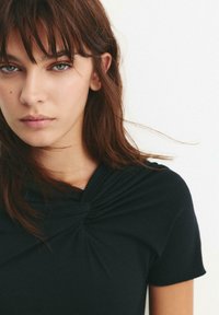 Sort top med korte ærmer og knude-detalje ved halskanten, glat stof og en figursyet pasform, der fremhæver skuldre og hals.