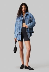 Chaqueta de mezclilla azul desvanecida con grandes bolsillos frontales, cierres de botón y un corte relajado, llevada sobre una blusa negra ajustada y pantalones cortos negros.