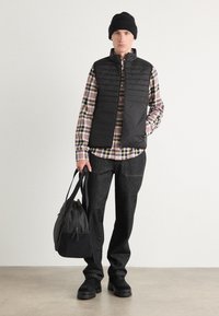 Colete preto almofadado sobre uma camisa xadrez, calças de ganga escuras, gorro preto e sapatos pretos. A carregar uma mala cinza com detalhes pretos.