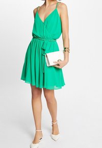 Grön ärmlös klänning med omlottdesign, insydd midja och fladdrig kjol. Kombinerad med en vit kuvertväska och beige klackar. Guldprydda remmar.