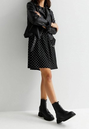 Kobieta w czarnej skórzanej kurtce, czarnej sukience w kropeczki i czarnych botkach na suwak, stoi na tle jasnego, jednolitego tła.