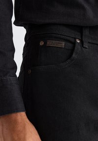 Close-up of a person wearing black denim jeans and a black long-sleeve shirt, focusing on the right hip pocket with metal rivets and a Wrangler label.