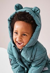 Veste matelassée en teal avec capuche ornée d'oreilles d'ours, fermeture éclair et patch étoile brodé sur la manche.