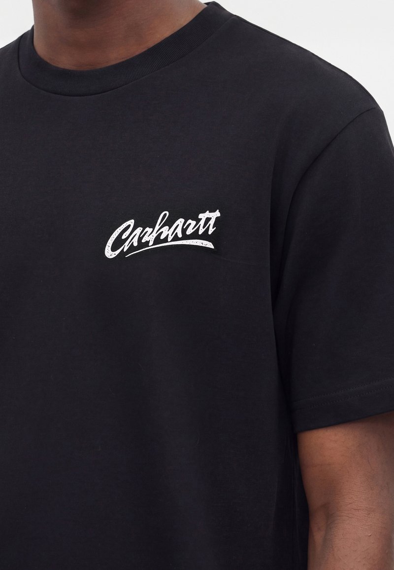 Person iført en sort T-shirt med korte ærmer og rund halsudskæring med hvidt "Carhartt"-logo på venstre bryst.