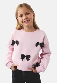 Różowy sweatshirt z czarnymi wstążkami przyszytymi na przodzie. Długie rękawy, okrągły dekolt, miękka tekstura. Noszony z ciemnymi spodniami.