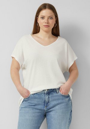 Frau mit langen, glatten Haaren, trägt Creolen, ein lockeres weißes T-Shirt mit V-Ausschnitt und hellblaue Jeans, Hände in den Taschen, neutraler Hintergrund.