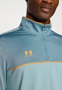 Top deportivo de media cremallera en tejido azul ligero con acentos naranjas. Presenta un patrón texturizado y el logo de Under Armour en el pecho.