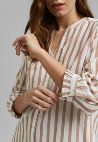 Blouse rayée en blanc et brun doux. Présente un col en V, une fermeture à boutons sur le devant et des poignets froncés. Fabriquée en tissu léger avec une coupe décontractée.