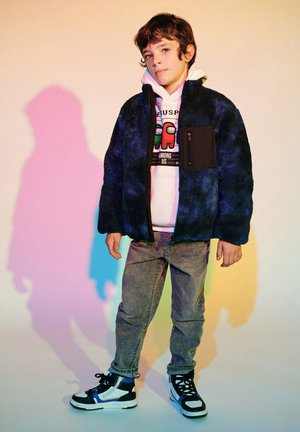 Giacca in pile blu navy con tasca nera, indossata sopra un hoodie bianco. Jeans grigi e sneaker nere completano l'outfit. Texture morbida evidente.