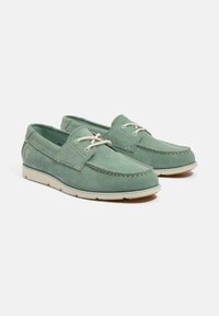 Scarpe da barca verde in suede con suola piatta color crema, punta rotonda, lacci abbinati e cuciture decorative lungo il bordo. Dettaglio del logo discreto sul lato.
