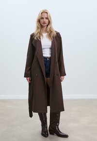Bruine trenchcoat met dubbellaagse knopen, brede kraag en lange mouwen, gecombineerd met een witte top, denim shorts en bruine kniehoge laarzen.