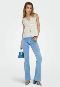 Witte mouwloze blouse met een oogjesdesign, strikdetail bij de taille, gecombineerd met lichtblauwe flared jeans en een bijpassende handtas.
