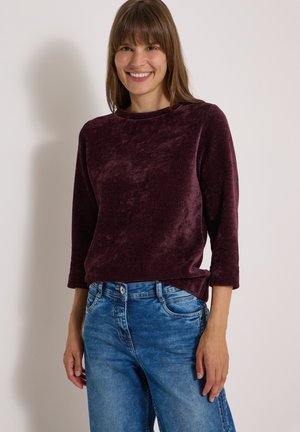 Femme souriante aux cheveux bruns lisses portant un pull violet foncé à manches longues et un jean bleu en denim sur un fond clair uni.