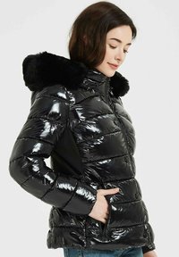 Veste en duvet noire avec finition brillante et matelassée, capuche bordée de fourrure et poches latérales. Présente un design ajusté et des poignets côtelés.