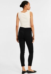 Mustat kapeat farkut, joissa on korkea vyötärö ja takataskut, yhdistettynä valkoiseen hihattomaan puseroon. Mallia kuvataan takaa. Tasainen kangas.