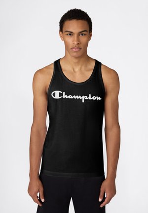 Hombre joven con una camiseta sin mangas negra con el logo blanco "Champion", posando de pie frente a un fondo blanco liso, mirando hacia adelante.