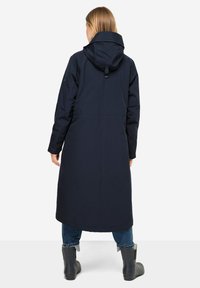 Parka lungo navy con cappuccio, caratterizzato da una vita sfiancata, texture liscia e tasche laterali. Indossato sopra jeans blu e stivali da pioggia neri.