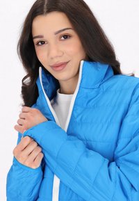 Jachetă puffer de culoare bleu deschis, cu guler înalt, fermoar frontal și manșete ribbate. Prezintă un design matlasat și un fermoar alb în contrast.