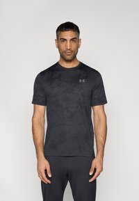 Μαύρο αθλητικό μπλουζάκι με κοντά μανίκια σε διακριτό υφασμάτινο μοτίβο, με στρογγυλή λαιμόκοψη και λογότυπο στο αριστερό μέρος του στήθους. Φορεμένο με σκούρο παντελόνι.