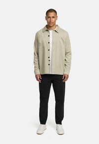 Light beige button-up shirt with two front pockets, worn over a white tee, paired with black pants and white sneakers.