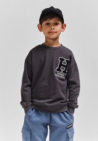 Szara bluza z dużym logo "H" i napisem "HUMMEL SPORTSWEAR", w zestawieniu z niebieskimi spodniami typu cargo z bocznymi kieszeniami i naszywką z logo.
