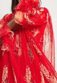 Punainen laskostettu mekko, jossa on kukkakuosi, röyhelöiset hihat ja läpikuultava kerrosrakenne, esittelee monimutkaista yksityiskohtaisuutta ja rohkeaa väri kontrastia.