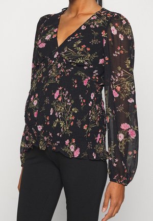 Femme portant un chemisier noir à manches longues avec des motifs floraux roses et verts et un pantalon noir ajusté sur un fond clair uni.