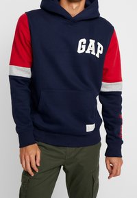 Felpa con cappuccio di un blu navy scuro con maniche accentate di rosso e grigio, con logo "GAP" sul petto e tasca frontale. Realizzata in morbido materiale felpato.