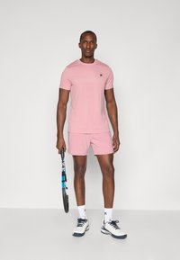 Light pink athletic t-shirt and matching shorts; fabric appears soft. Model holds a tennis racket, wearing white and navy sneakers with white socks.