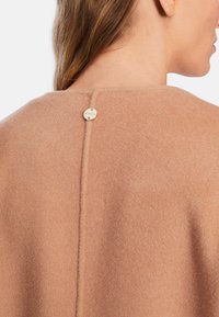 Beige Wollmischungsteil mit glatter Textur, das über einen Knopfverschluss hinten verfügt und eine vertikale Naht in der Mitte hat.