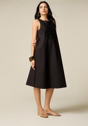 Femme en robe midi noire sans manches à broderies florales, boucles d’oreilles anneaux dorées, bracelets marron et sandales plates dorées, debout sur un fond uni.