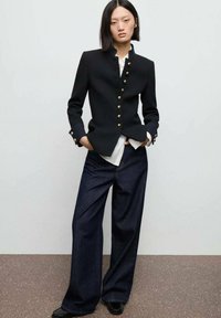 Černý saka na míru se zlatými knoflíky v kombinaci s volnými modrými džíny; hladká tkanina, strukturovaný design a kontrastní vrstvy.