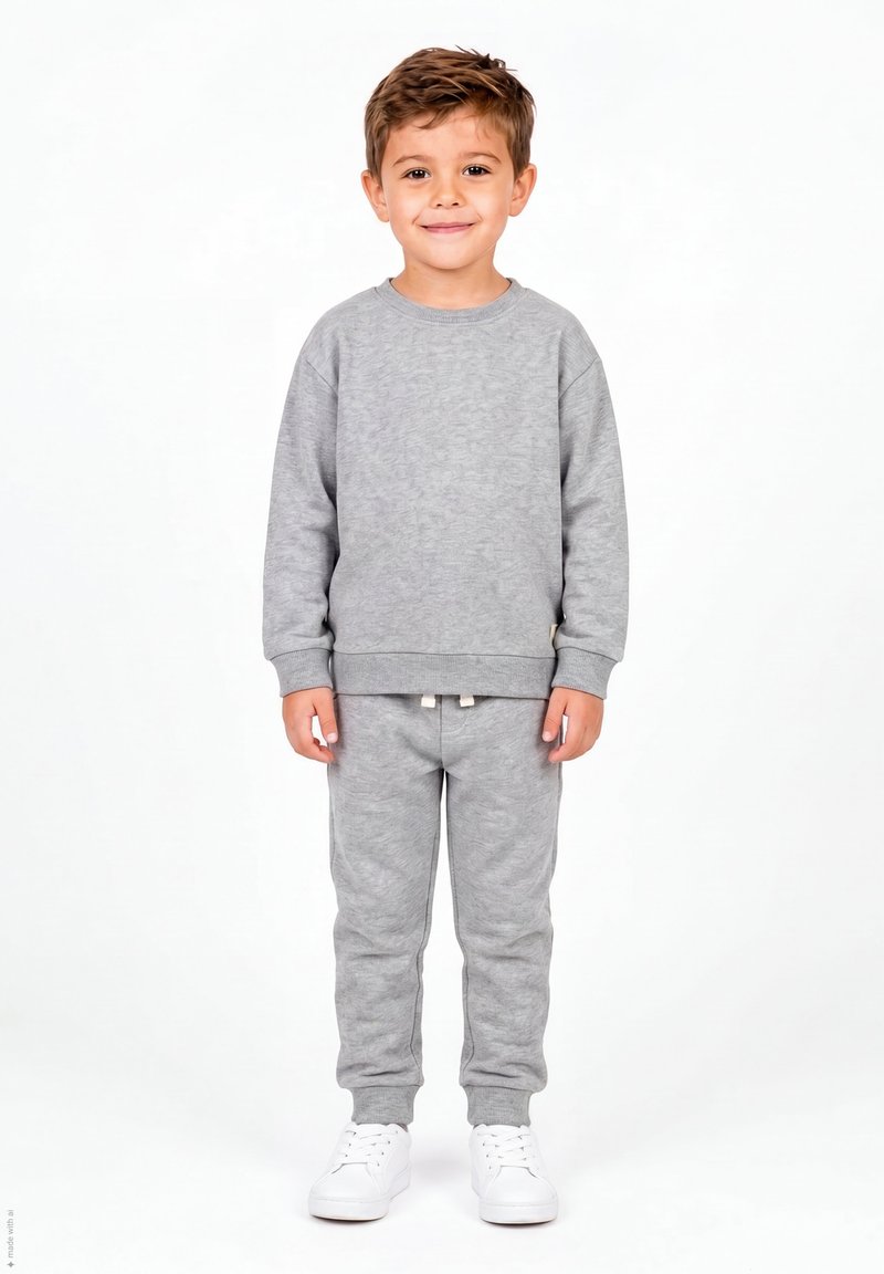 Jeune garçon debout et souriant, portant un sweat-shirt gris et un pantalon de survêtement avec des baskets blanches, sur fond blanc uni.
