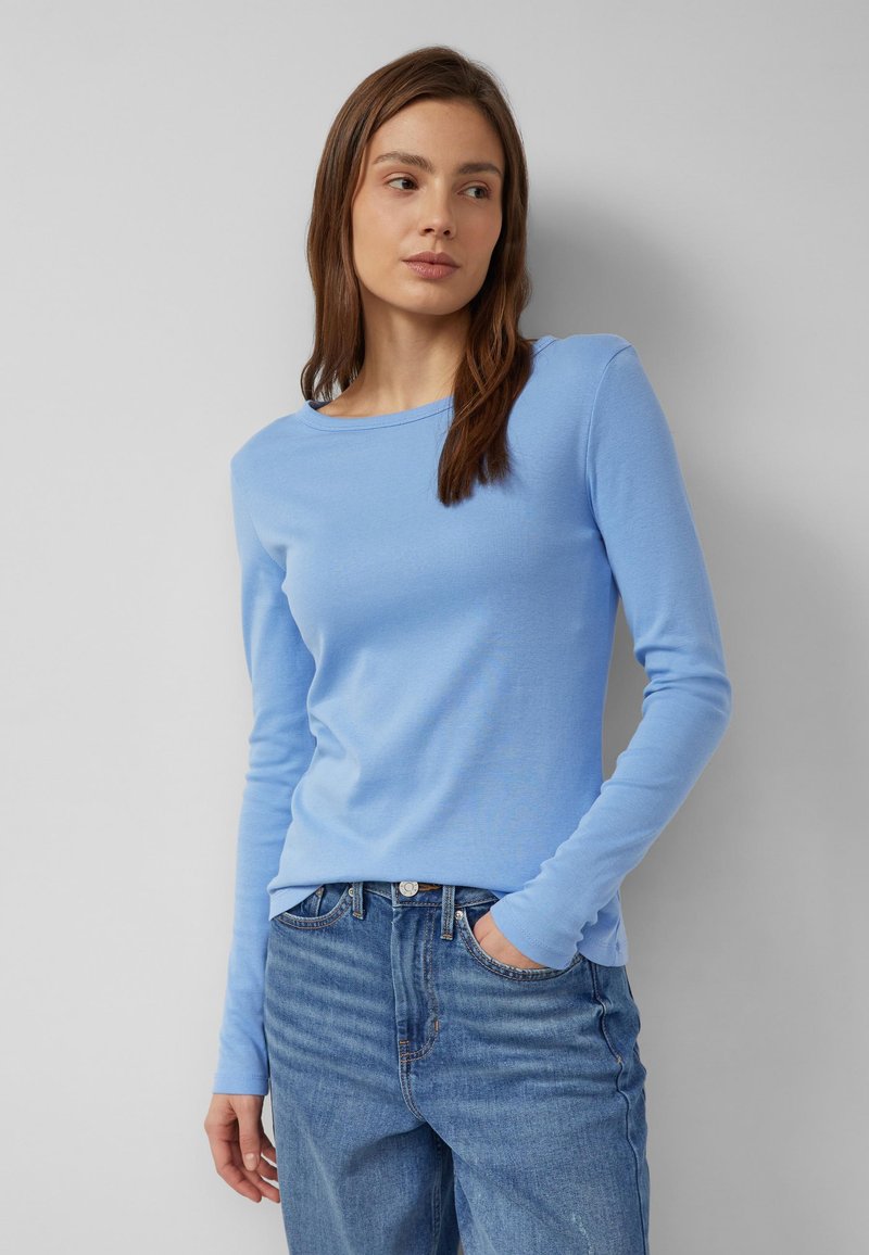 Mujer con cabello largo y castaño, vestida con una camisa de manga larga azul claro y jeans azules, de pie con una mano en el bolsillo frente a un fondo gris.