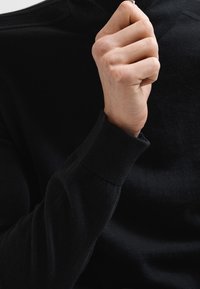 Czarny sweter z golfem o teksturowanej tkaninie, dopasowanych rękawach i wysokim dekolcie; dłoń chwytająca kołnierz, ukazująca szczegóły tkaniny.