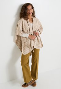 Poncho beige à motifs avec une ceinture à nouer, porté sur un haut blanc, associé à un pantalon vert olive et des chaussures marron à imprimé léopard.