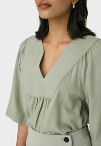 Helles grünes Blusenshirt mit puffierten Ärmeln, V-Ausschnitt und Raffung am Bust. Weicher, strukturierter Stoff mit leichtem Glanz.