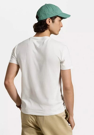 T-shirt bianco di cotone a maniche corte, indossato al contrario. Un cappellino verde menta si trova sopra, abbinato a pantaloni beige. Texture morbida visibile.