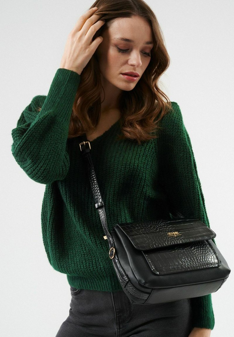 Czarna torba na ramię z teksturowanym wzorem krokodyla, złotymi detalami i regulowanym paskiem, zestawiona z zielonym swetrem dzianinowym.