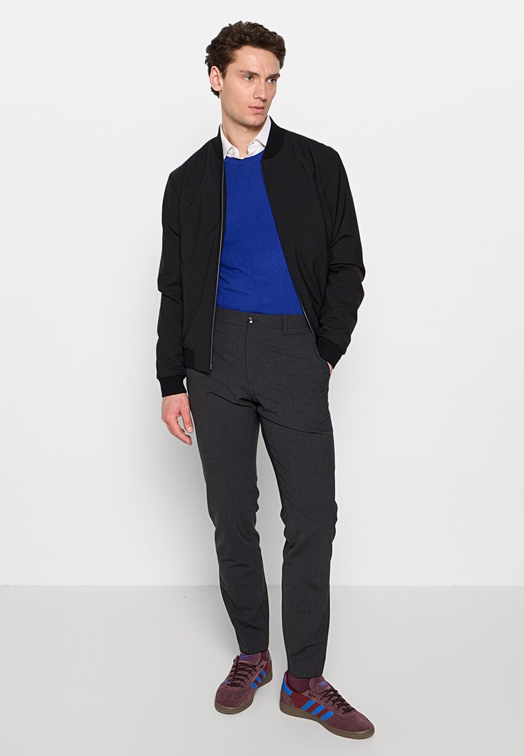 Schwarze Bomberjacke über einem blauen Sweater, graue Slim-Fit-Hose und bordeauxfarbene Sneakers mit blauen Streifen, präsentiert an einem stehenden männlichen Model.