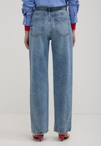 Jeans a vita alta blu chiaro a gamba larga con una texture sbiadita, dotati di due tasche posteriori e orli finiti. Indossati con sandali rossi con tacco.