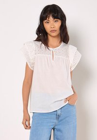 Hvid bluse med broderet øjenlågdesign på korte flæseærmer, nøglehuldetalje ved halsudskæringen og en afslappet pasform. Bruges med blå jeans.