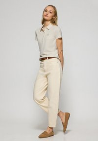 Polo beige con colletto e tre bottoni, abbinato a pantaloni a vita alta color avorio e scarpe slip-on marrone chiaro.