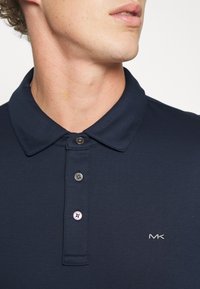 Mørkeblå polo-skjorte med krage, som har tre sølvknapper og en brodert "MK"-logo på venstre bryst. Myk tekstur.