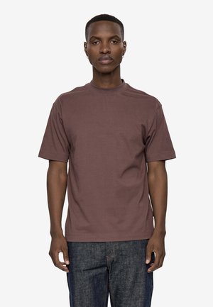Mand stående, vendt fremad iført en ensfarvet brun kortærmet t-shirt og mørkeblå jeans mod en hvid baggrund.