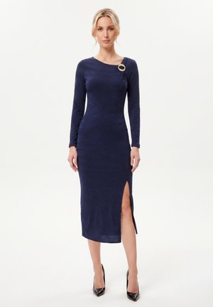 Femme portant une robe midi bleu marine à manches longues avec une fente latérale et une broche décorative près du col, associée à des talons noirs.