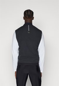 Schwarze ärmellose Weste mit hohem Kragen, strukturiertem Stoff und reflektierendem Streifen auf der Rückseite. Über einem weißen Langarmshirt getragen.