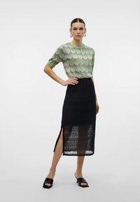 Top tricotat cu model în zigzag verde și maro, cu mâneci scurte, asortat cu o fustă lungă neagră, texturată, cu o desfăcutură pe lateral. Sandale negre tip slip-on.