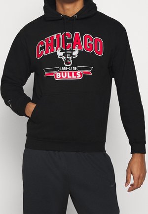 Personne portant un sweat à capuche noir avec le logo et le texte Chicago Bulls en rouge et blanc, associé à un pantalon de sport foncé.