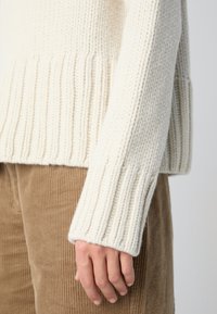 Gros plan sur la main d'une personne portant un pull en maille épaisse couleur crème et un pantalon en velours côtelé marron, sur un fond uni.